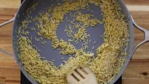 Toasting orzo pasta in a skillet with a wooden spatula on a wooden surface.