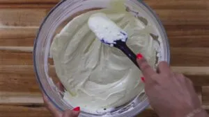 Mixing creamy cake batter in a glass bowl with a spatula on a wood surface.