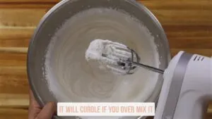 Electric mixer whipping cream in a bowl with a note saying, It will curdle if you over mix it.
