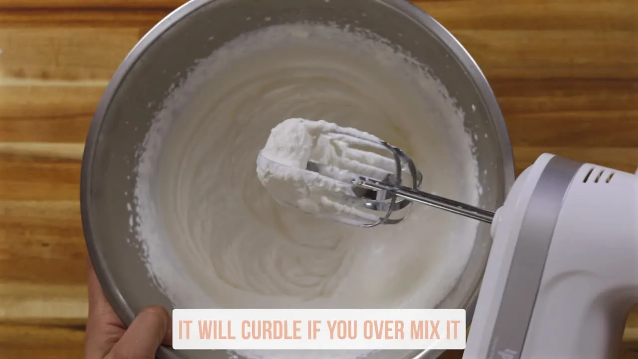 Electric mixer whipping cream in a bowl with a note saying, It will curdle if you over mix it.