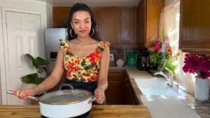 Woman in floral top holding white pot, standing in a cozy kitchen with wooden cabinets and vibrant flowers.