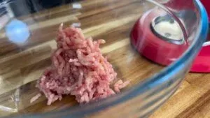 Ground meat in a glass bowl on a wooden counter beside a red kitchen appliance.