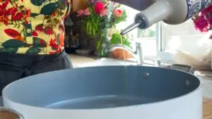 Pouring olive oil into a frying pan in a bright kitchen with floral decor in the background.