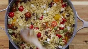 Skillet filled with orzo pasta, cherry tomatoes, olives, and feta cheese on a wooden surface.