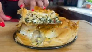 Person cutting a slice of crispy, flaky spinach and cheese pie on a wooden board.