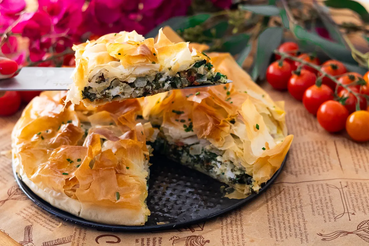Freshly baked spinach and feta pie with golden phyllo crust, served on a plate, cherry tomatoes in background.