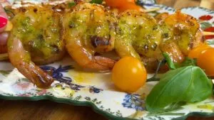Grilled shrimp with pesto glaze on skewer, served with cherry tomatoes and basil on a floral plate.