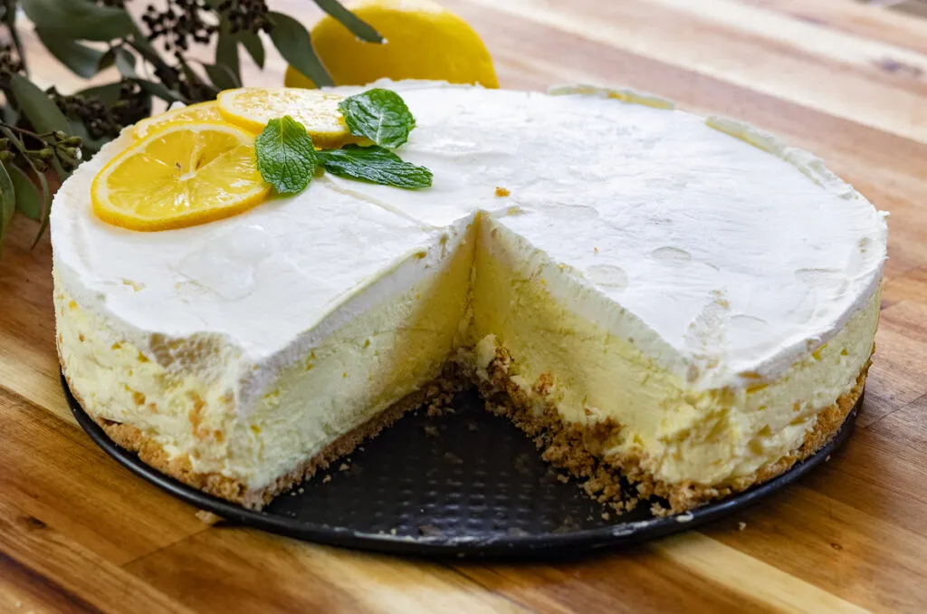 No-Bake Mascarpone Lemon Cheesecake with a slice missing, garnished with fresh lemon slices and mint on a wooden table.