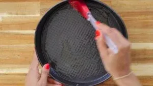 Greasing a round baking pan with oil using a red silicone brush on a wooden surface.