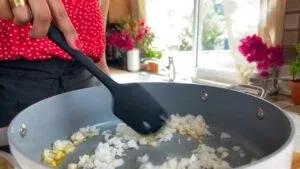 Person in a red polka dot shirt cooking chopped onions in a pan with a black spatula, bright kitchen setting.