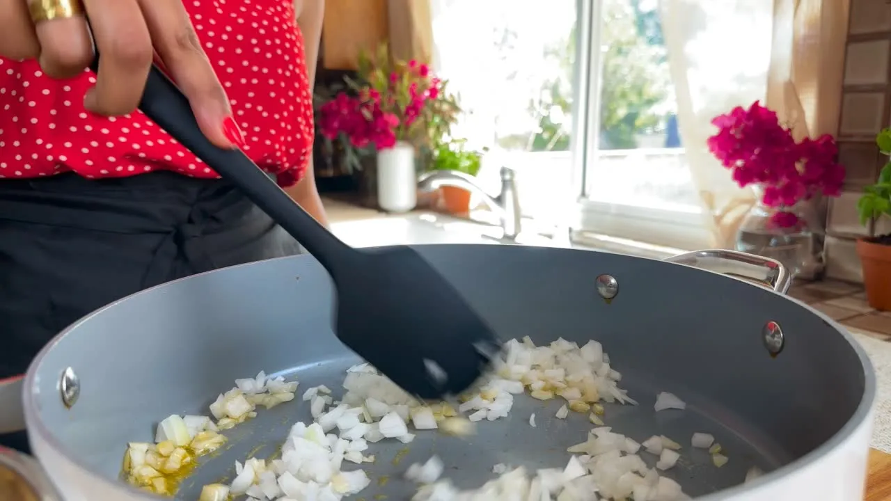 Person in a red polka dot shirt cooking chopped onions in a pan with a black spatula, bright kitchen setting.