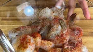 Raw shrimp seasoned with paprika and pepper in a glass bowl on a wooden surface.