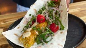 Plate with grilled meat wrap, fresh greens, cherry tomatoes, and sauce on a wooden table, ready to eat.
