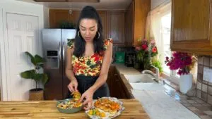 Woman in colorful dress prepares a meal in a homey kitchen with flowers and wooden cabinets.