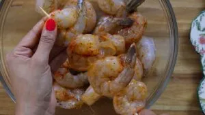 Person holding skewer with seasoned shrimp, ready for grilling in kitchen.