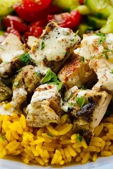 Grilled chicken with yellow rice, creamy sauce, and fresh cucumber-tomato salad on a white plate.