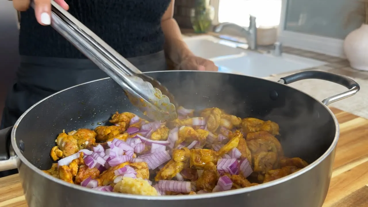 Person cooking chicken shawarma with onions in a pan using tongs in a kitchen.