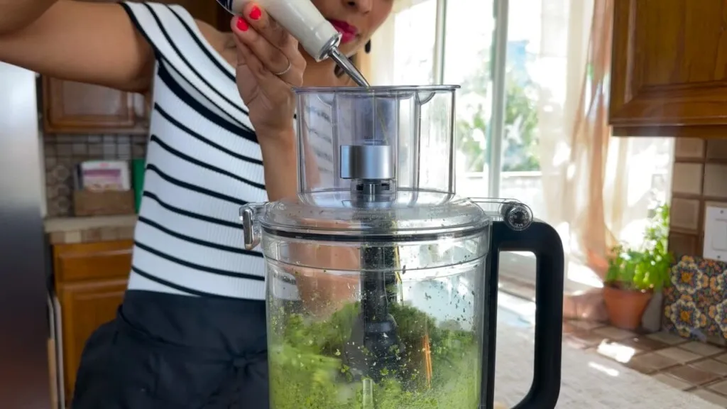 Woman making pesto using a food processor in a bright kitchen.