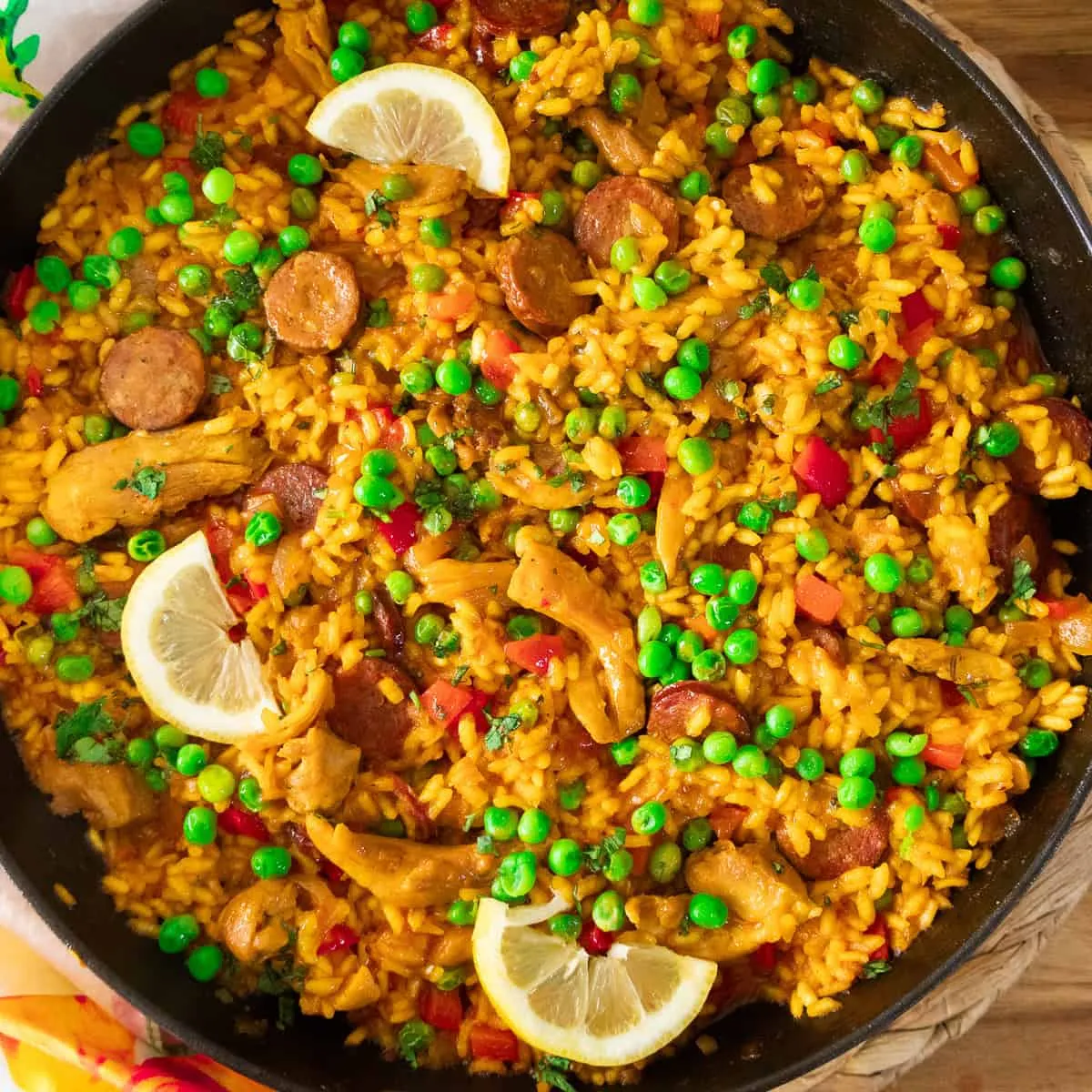 Pan of vibrant paella with saffron rice, peas, chorizo, chicken, red peppers, and lemon wedges.