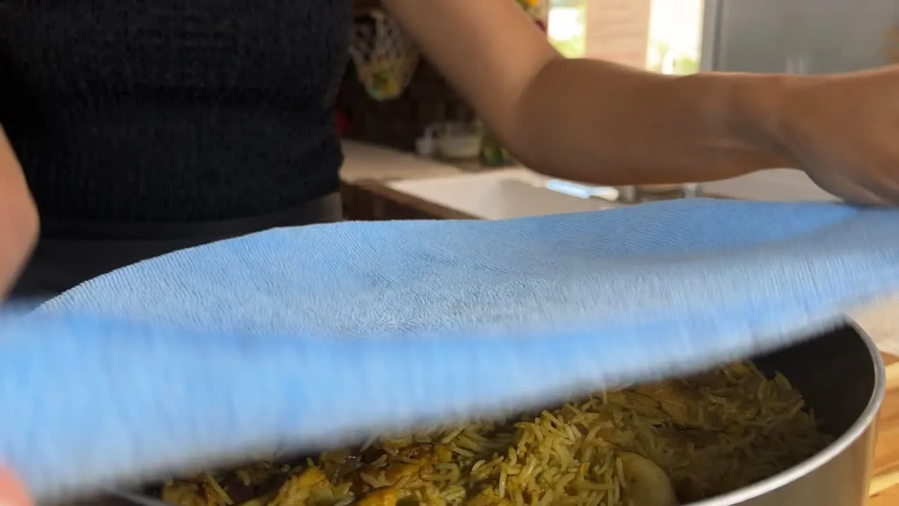 Person covering a pot of fragrant rice with a blue cloth in a kitchen setting.