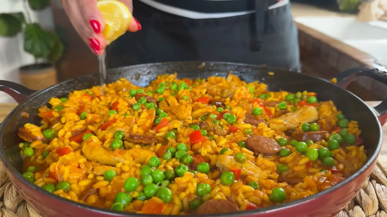 Person squeezing lemon over colorful paella with chicken, sausage, peas, and red peppers in a large pan.