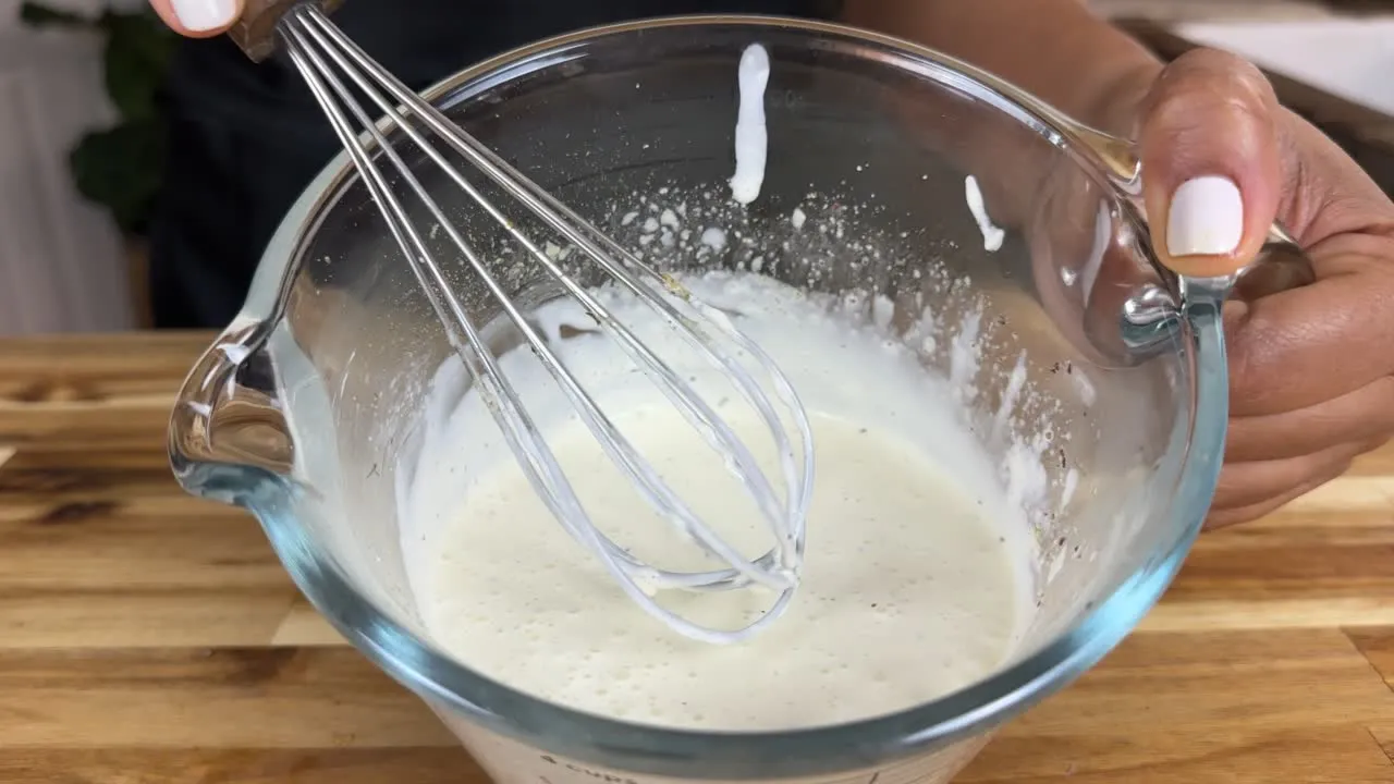 Person whisking a white sauce in a glass bowl on a wooden table.