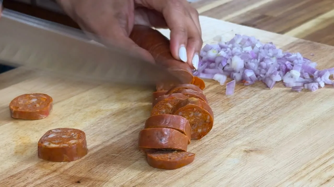 Hands slicing sausage on a cutting board next to finely chopped onions.