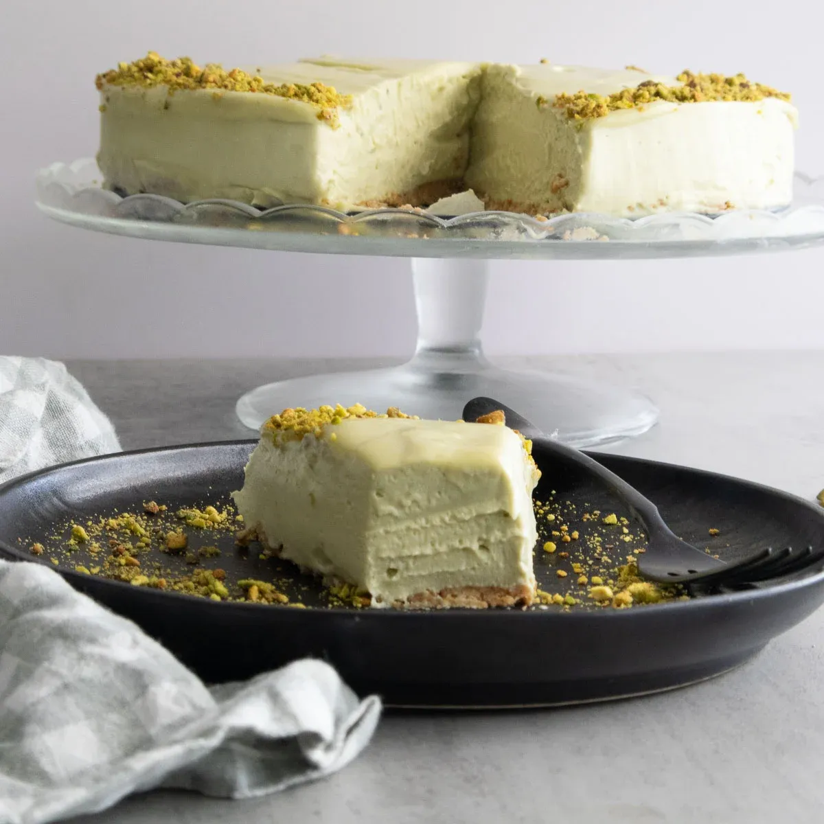 Slice of no bake mascarpone pistachio cheesecake on black plate, garnished with crushed pistachios, on a cake stand.