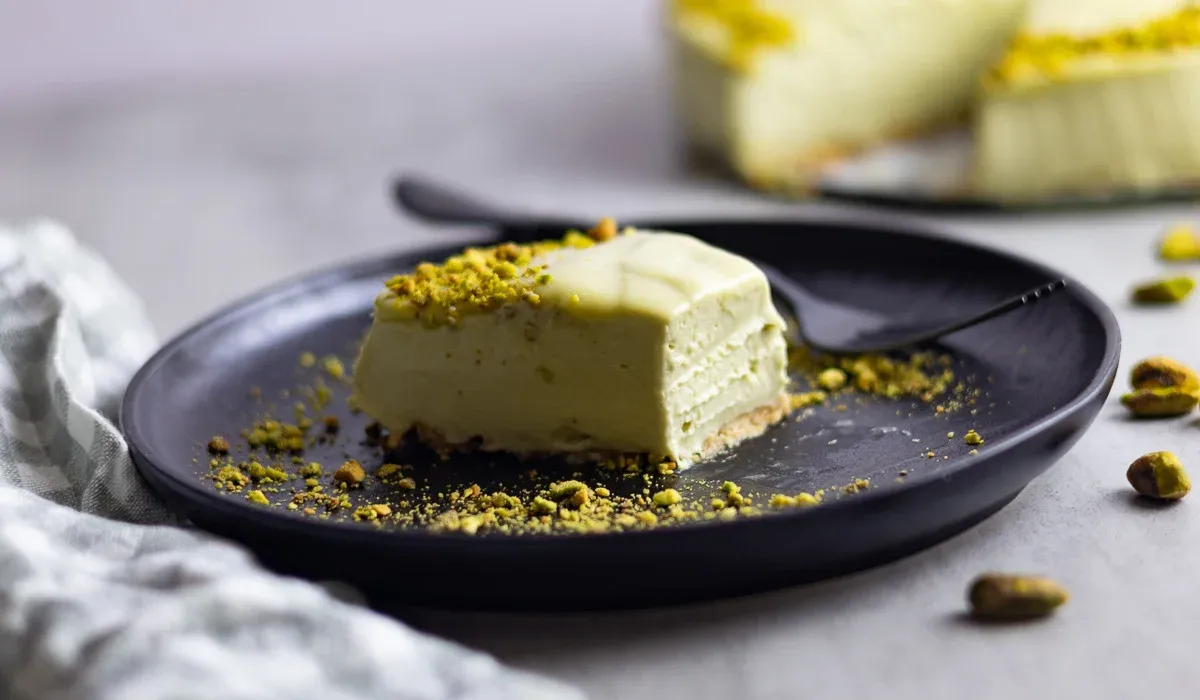 Creamy pistachio cheesecake slice on a black plate, with crushed pistachios on top and around.