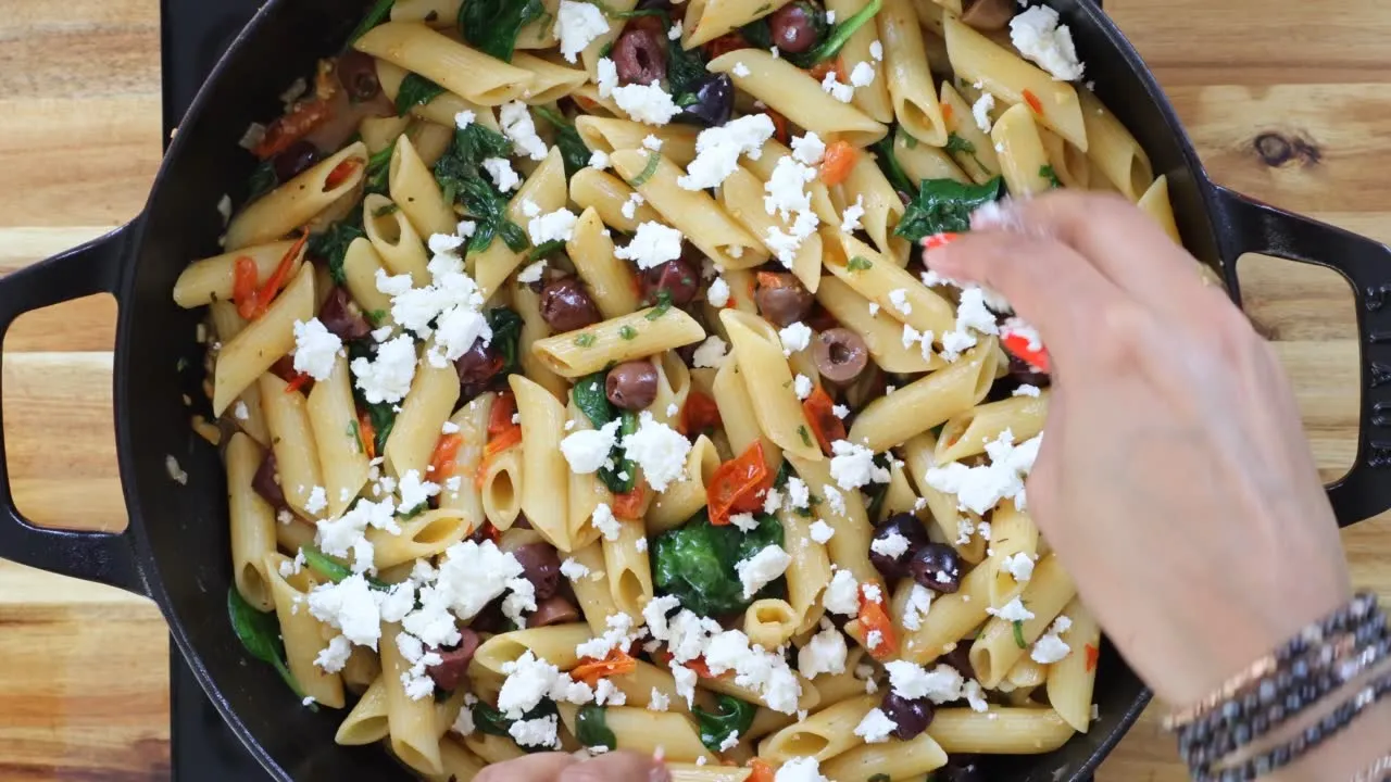 Penne pasta in a skillet with spinach, olives, tomatoes, and feta cheese being mixed by hand.