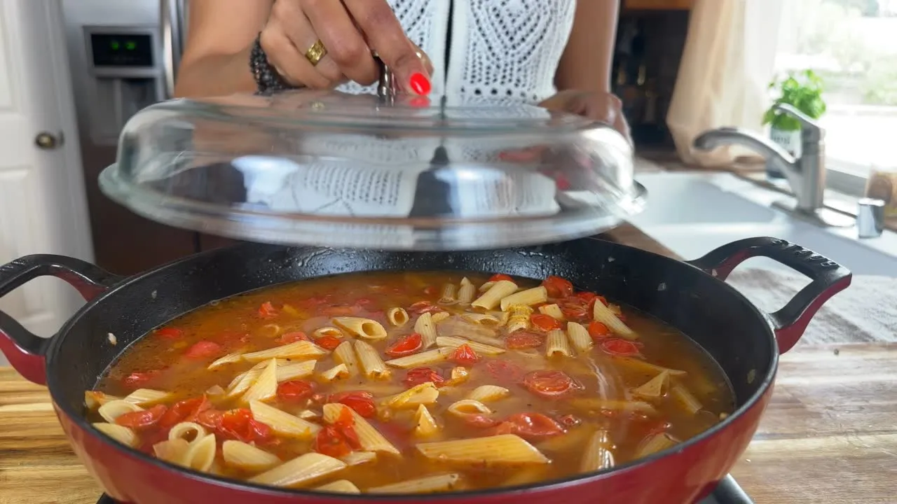 Cooking penne pasta with tomatoes in a skillet, person lifting lid in a bright kitchen setting.