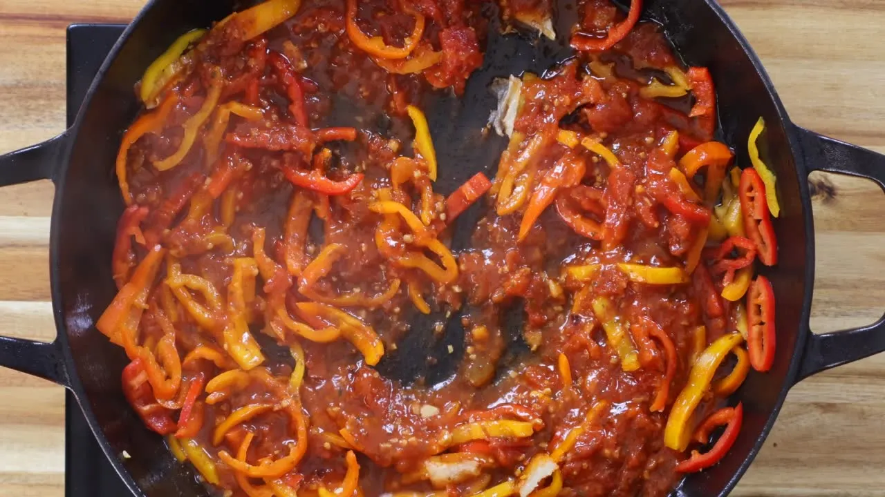 Sautéed red and yellow bell peppers in a cast iron skillet with tomato sauce on a wooden table.