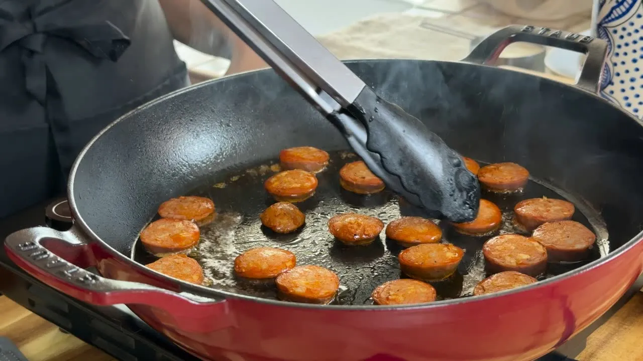 Chef cooking sliced sausage in a red pan with tongs, capturing the cooking process.