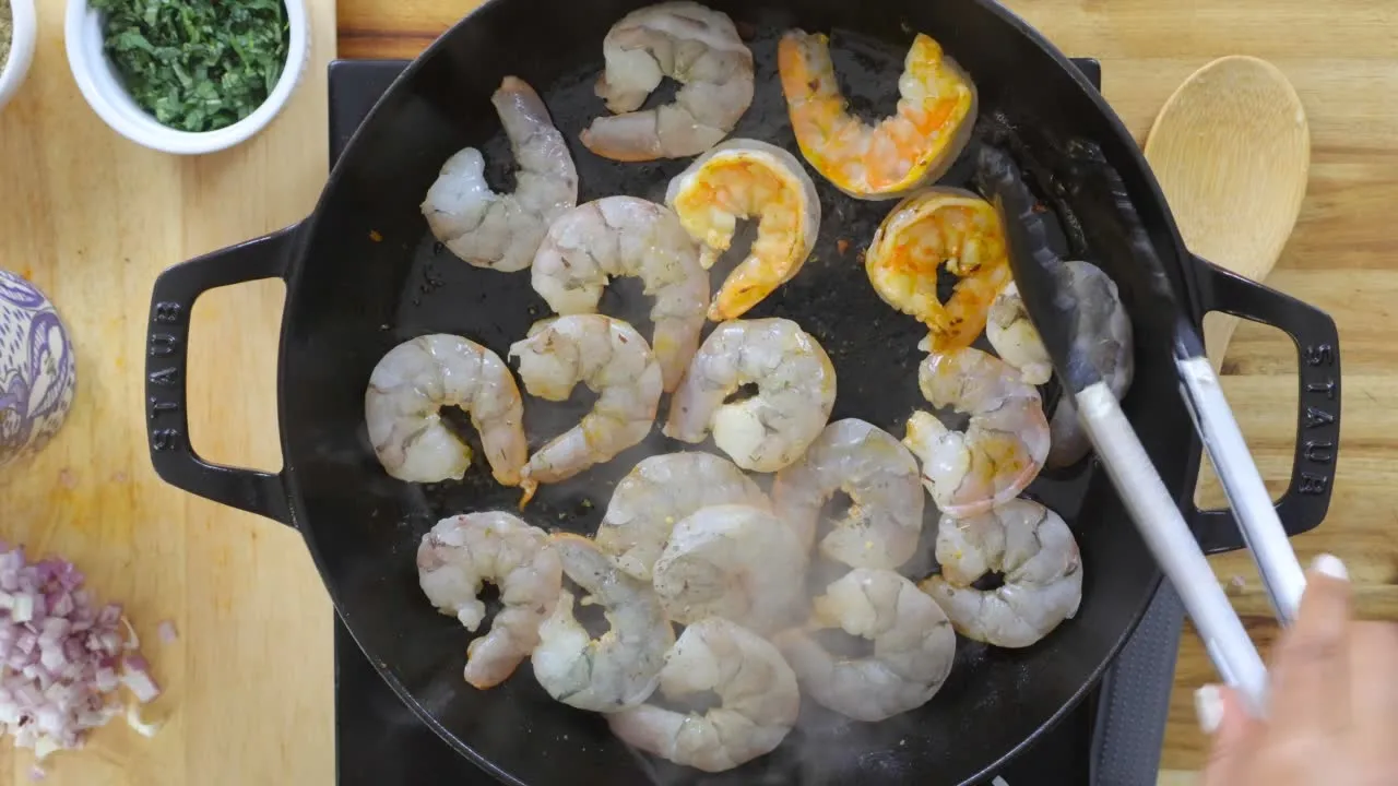 Raw and cooked shrimp in a skillet with tongs, herbs, and shallots on a wooden countertop.