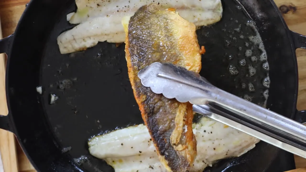 Grilled fish fillet being flipped with tongs in a hot cast iron skillet, sizzling and seasoned.