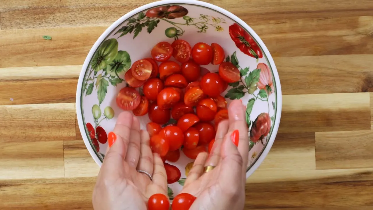 Hands holding fresh cherry tomatoes in a floral bowl on a wooden countertop.