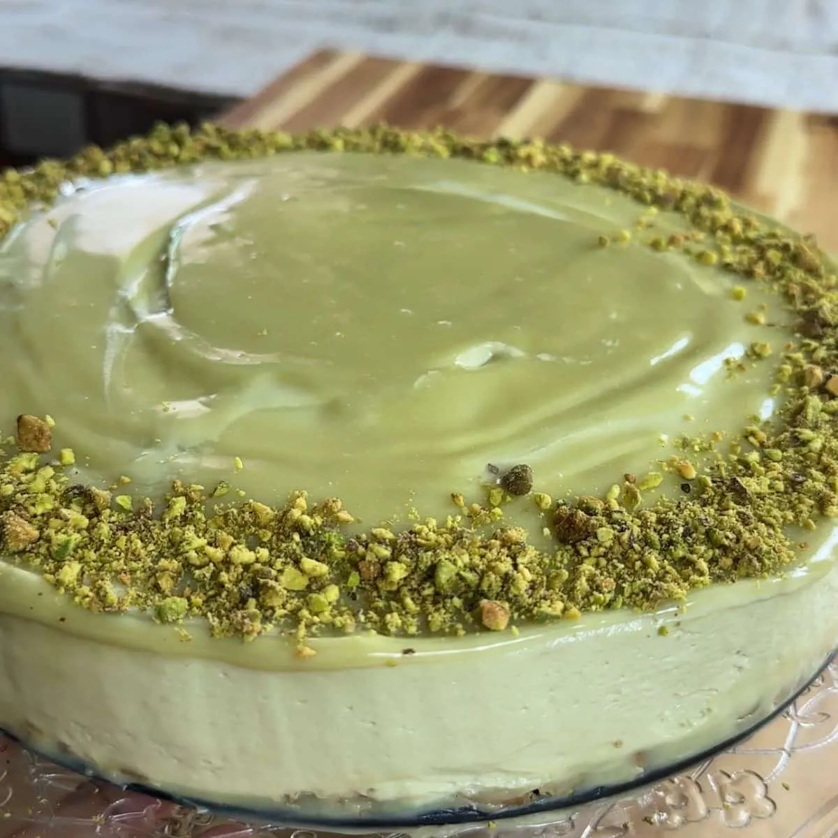 Creamy no bake pistachio cheesecake with a smooth green frosting and crushed pistachio topping on a wooden board.
