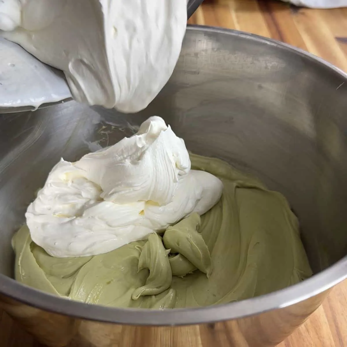 Mixing creamy white frosting with pistachio icing in a metal bowl on a wooden countertop.