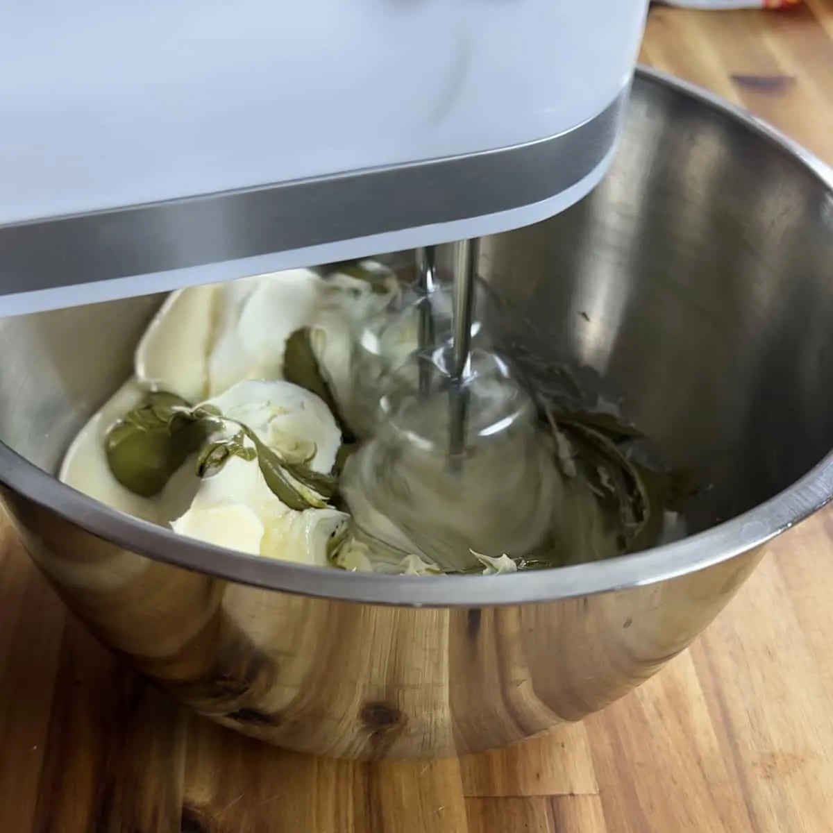 Electric mixer blending cream cheese and pickles in a metal bowl on a wooden countertop.