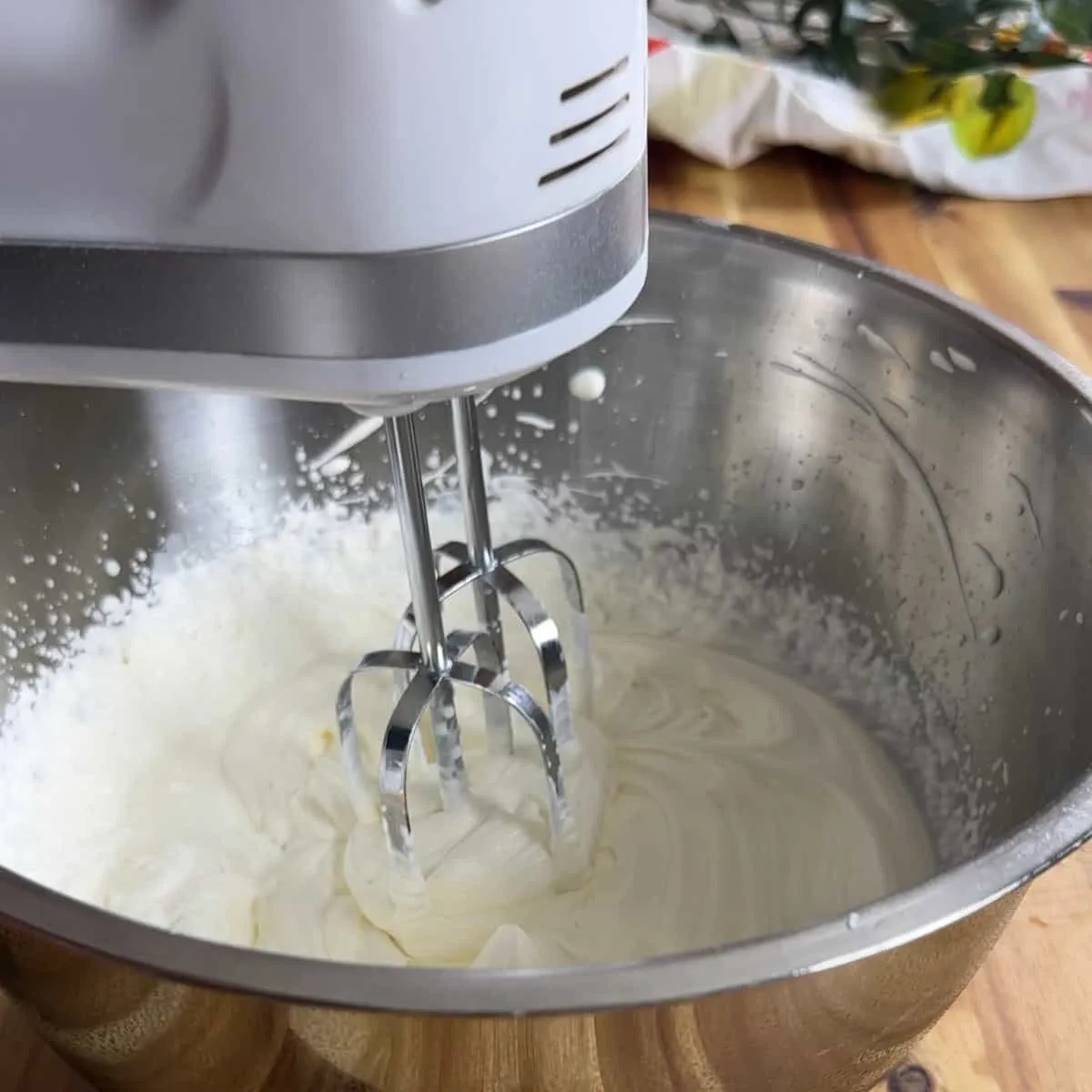 Electric mixer whipping cream in a stainless steel bowl on a wooden table.