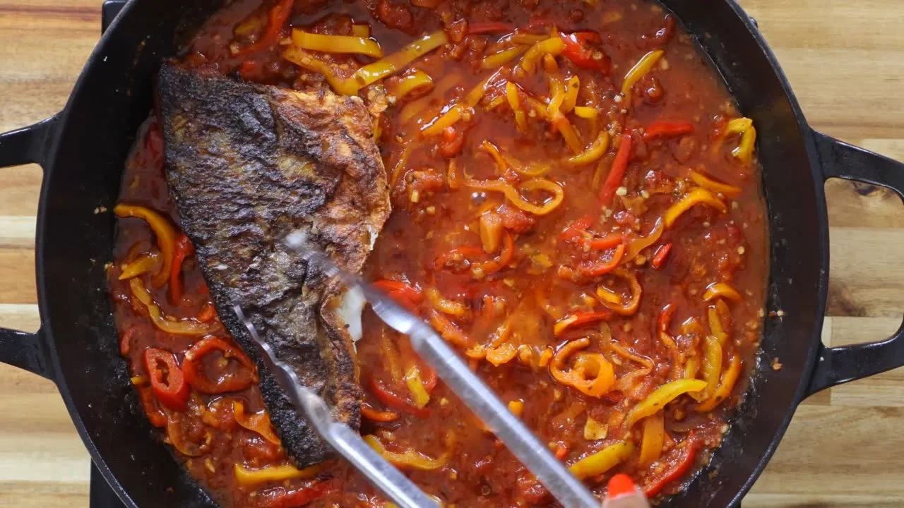 Fried fish in a pan with red and yellow bell peppers simmering in tomato sauce.