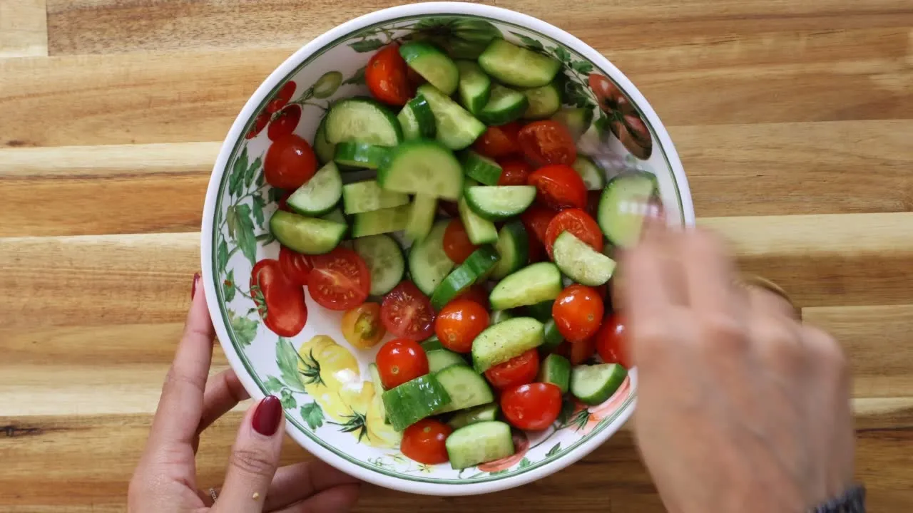 Fresh cucumber and cherry tomato salad being mixed in a decorative bowl on a wooden table.