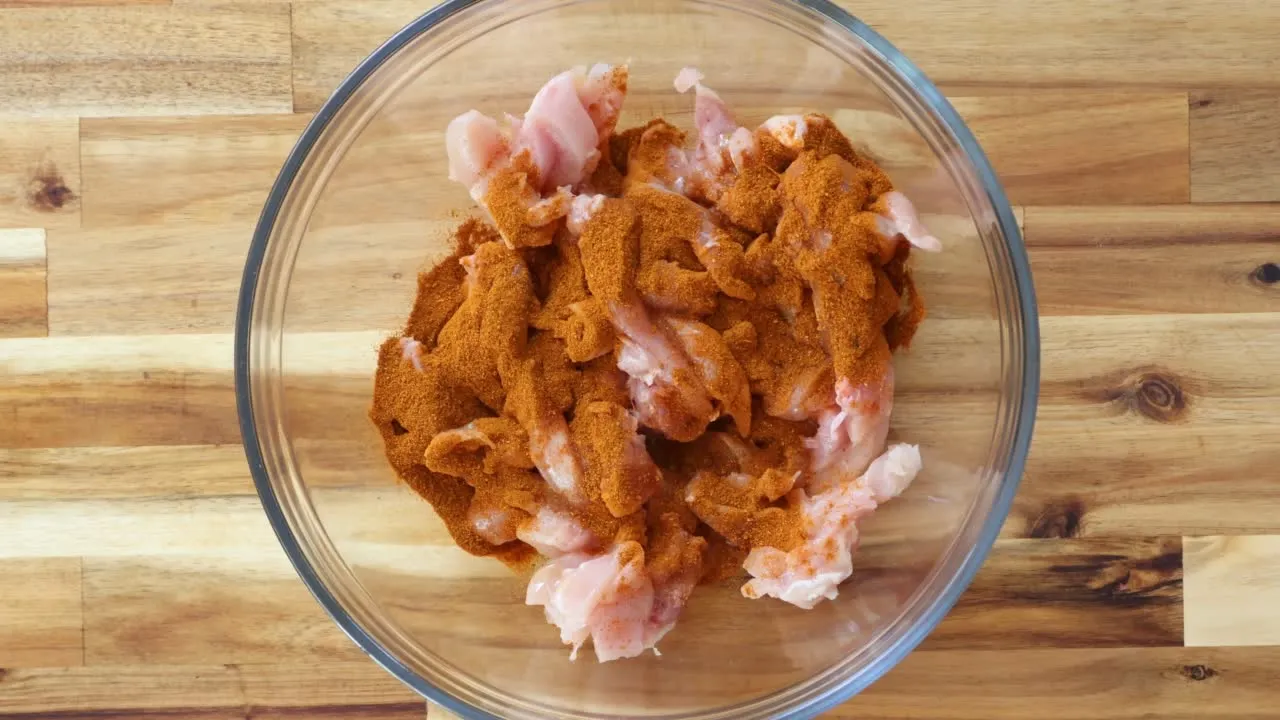 Raw chicken pieces seasoned with spices in a glass bowl, ready for cooking on a wooden countertop.