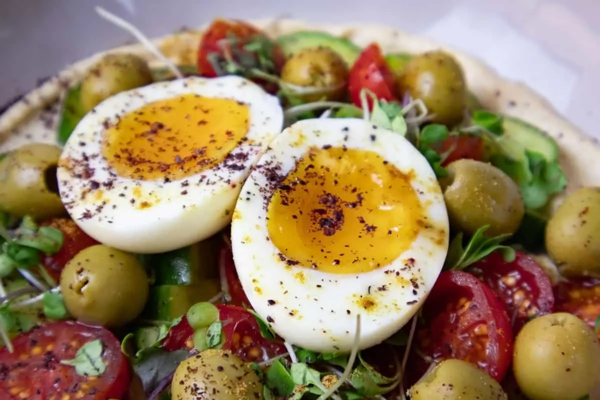 Mediterranean Hummus Bowl with Soft-boiled eggs on a vibrant salad with tomatoes, olives, and microgreens, sprinkled with spices.