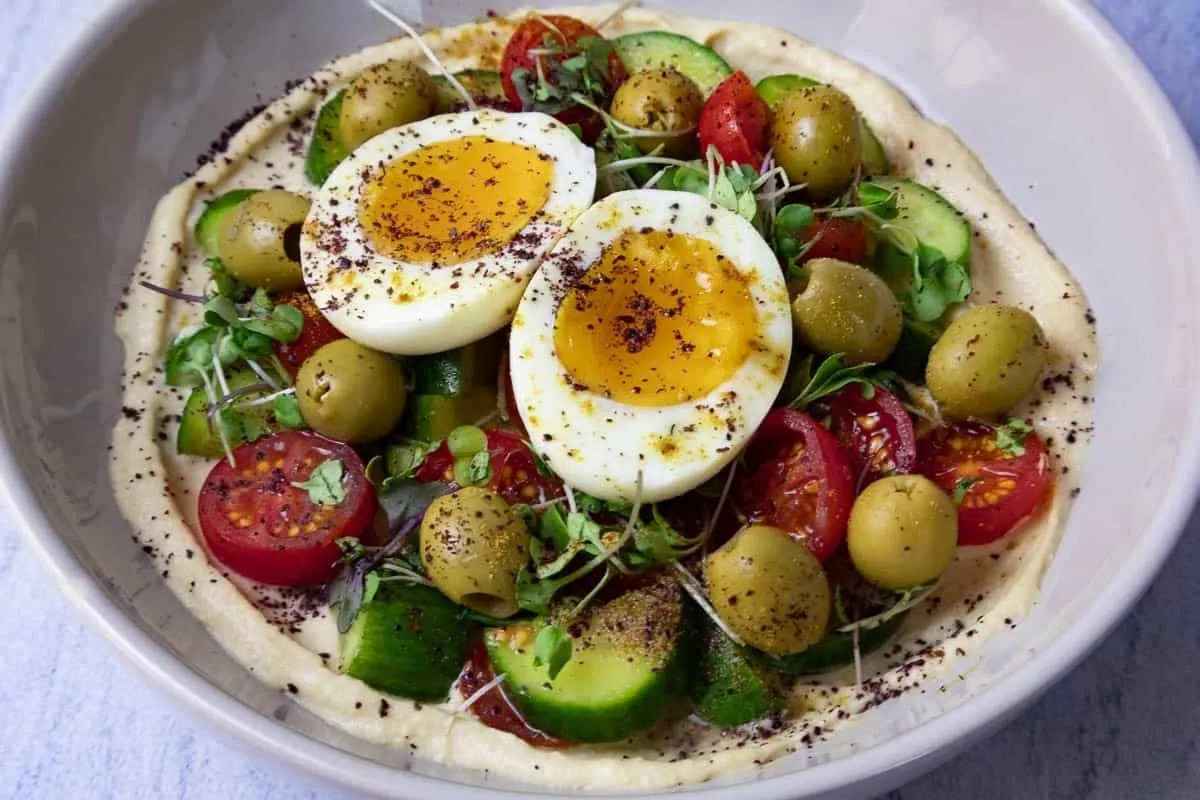 Mediterranean Hummus Bowl topped with soft-boiled eggs, cherry tomatoes, olives, and greens.