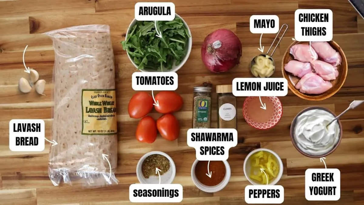 Ingredients for a shawarma wrap recipe on a wooden surface: lavash, chicken, spices, Greek yogurt, veggies, and more.
