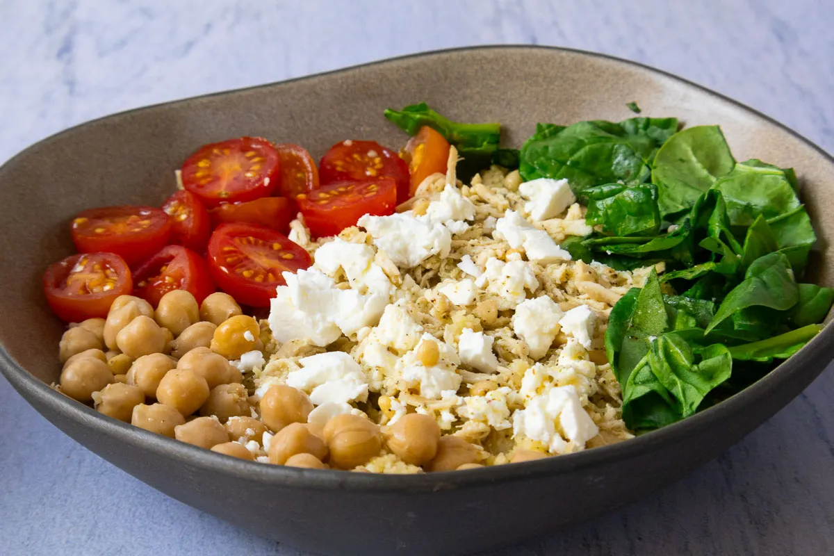 Bowl with chickpeas, cherry tomatoes, feta, spinach, and grains on a stone surface.