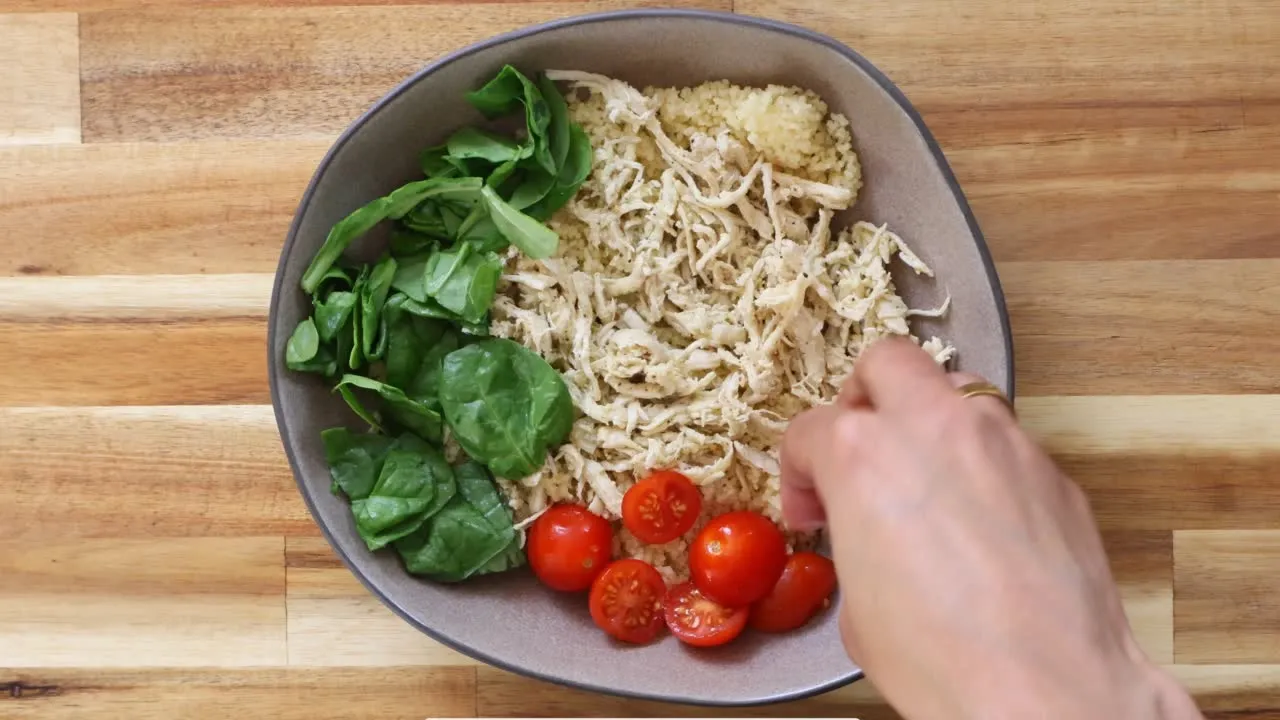 Bowl of shredded chicken, fresh spinach, cherry tomatoes, and couscous, with a hand arranging ingredients.