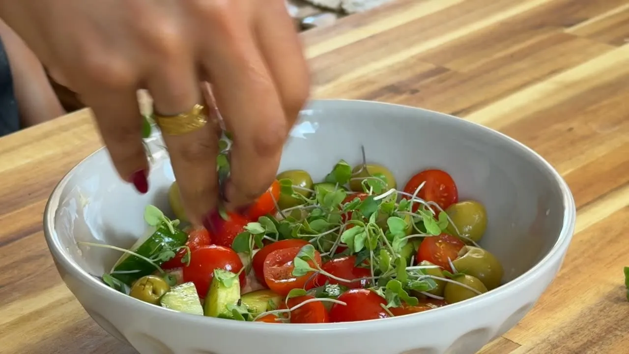 Hand preparing fresh cherry tomato salad with microgreens in a white bowl on wooden table.