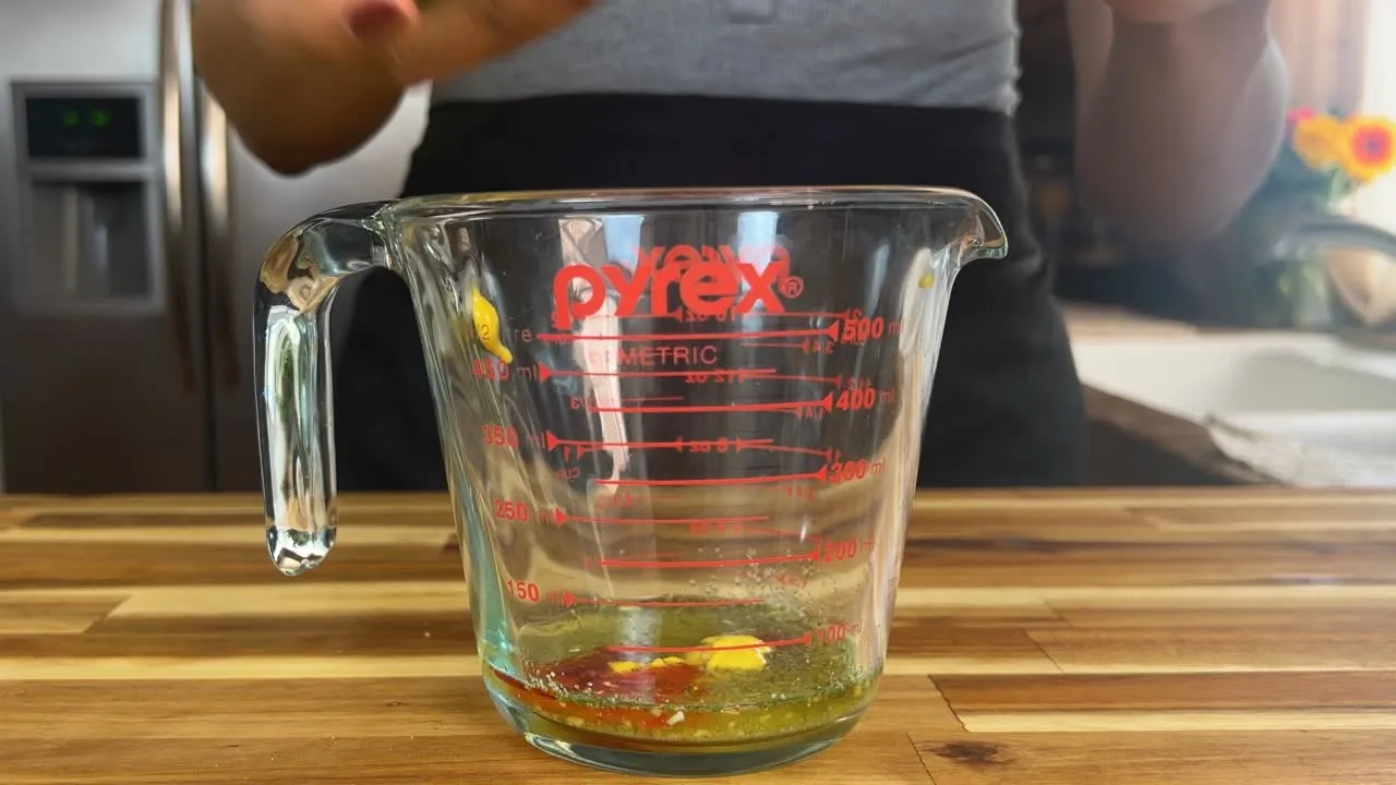 Glass measuring cup on wooden counter with oil and seasoning, kitchen in background.